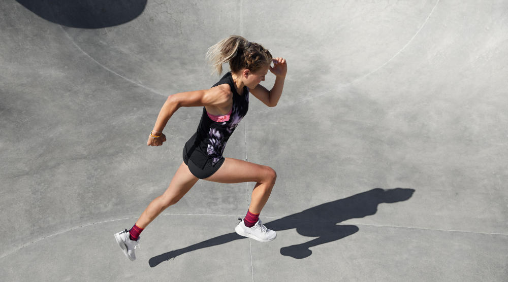 Läuferin mit Sprunggelenkbandagen im sommerlichen Outfit sprintet über Betonfläche	 Perspektive von seitlich oben