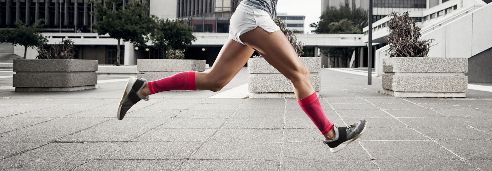 Frau mit pinken Wadensleeves läuft schnell und mit Beinen in der Luft durch eine urbane Betonwüste