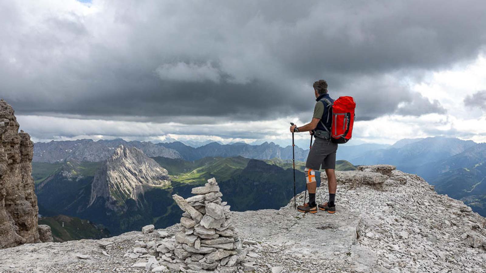 Wanderer auf einem Felsvorsprung mit Wander Kniebandage Eine Person mit einem roten Rucksack steht auf einem Felsvorsprung in den Dolomiten und blickt in die Ferne. Der Himmel ist bewölkt und es gibt eine Steinmännchen-Konstruktion im Vordergrund.