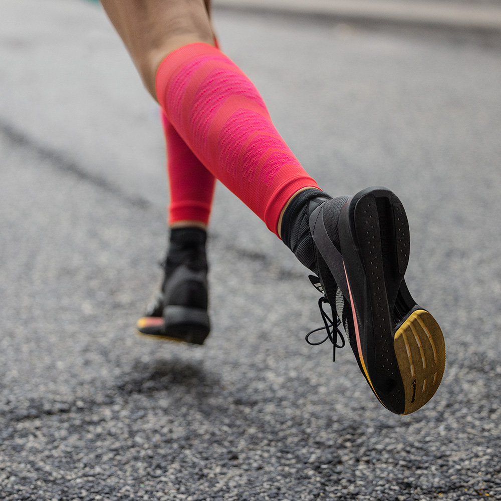 Detailaufnahme einer Läuferin auf Asphalt, die dynamisch nach vorne läuft. Sie trägt schwarze Laufschuhe und auffällige rote Run Performance Sleeves mit Kompression, die ihre Wadenmuskulatur unterstützen.