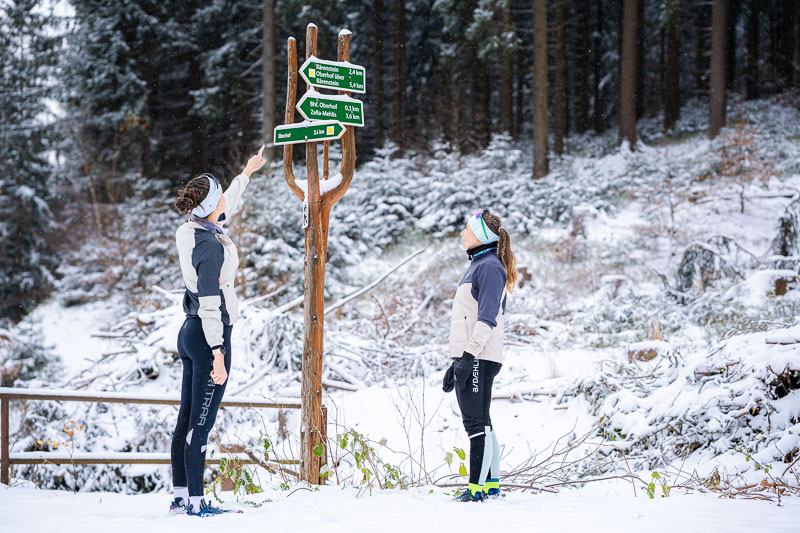Die richtige Route finden, im Winter richtig wichtig! Zwei Frauen in Winterlaufkleidung stehen vor einem hölzernen Wegweiser im verschneiten Wald. Eine der Frauen zeigt mit dem Finger auf die Schilder, während die andere aufmerksam schaut. Der Wegweiser zeigt verschiedene Wanderrouten.