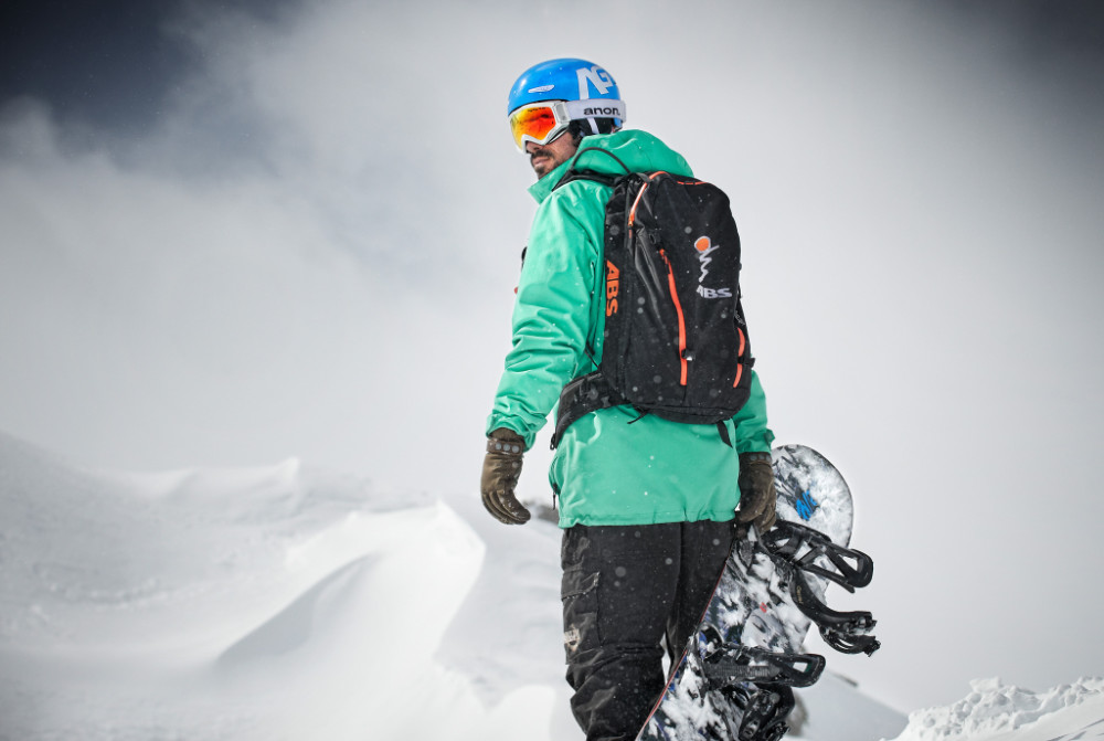 Snowboarder mit grüner Jacke und Board in der Hand blickt über seine Schulter nach hinten
