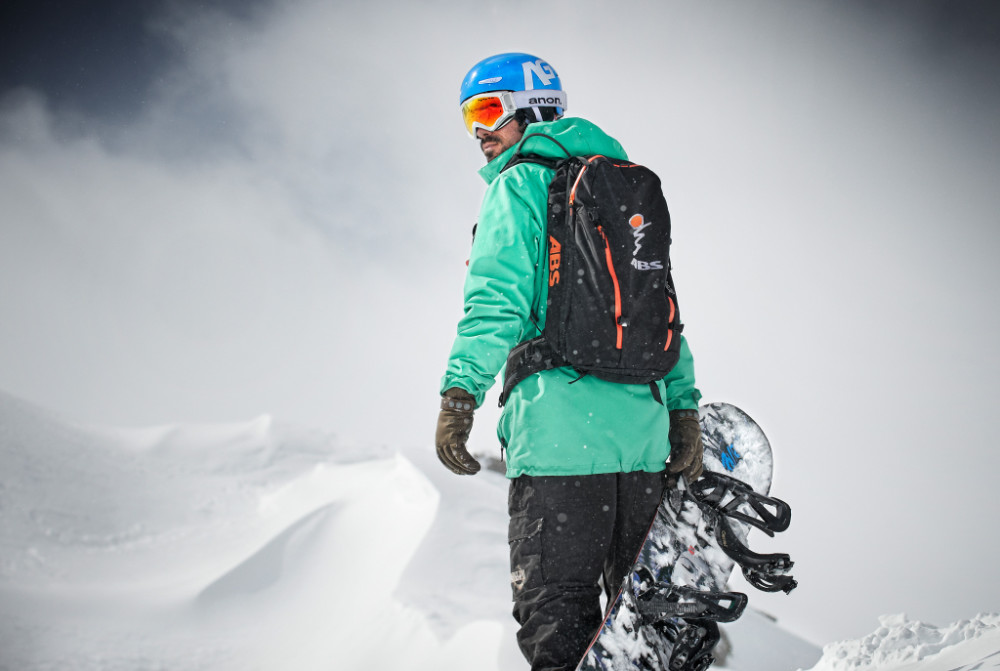 Was tun für kraftvolle Oberschenkel beim Snowboarden? Wie stärke ich meinen Rücken? Wir haben ein paar Tipps Snowboarder mit grüner Jacke und Board in der Hand blickt über seine Schulter nach hinten