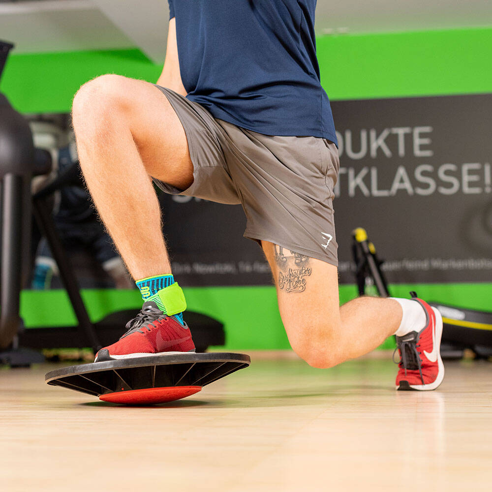 Mann in einem Fitnessstudio macht einen Lunge mit dem vorderen Bein auf einem Balance Pad stehend und trägt eine Sprunggelenkbandage