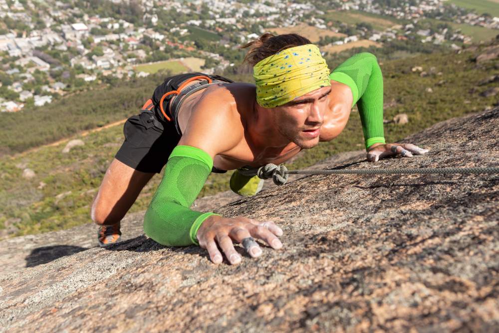 Durch Seil gesicherter Kletterer am Berg	 freier Oberkörper	 Bandana am Kopf und grüne Arm Sleeves	 im Tal ist eine Stadt zu erkennen