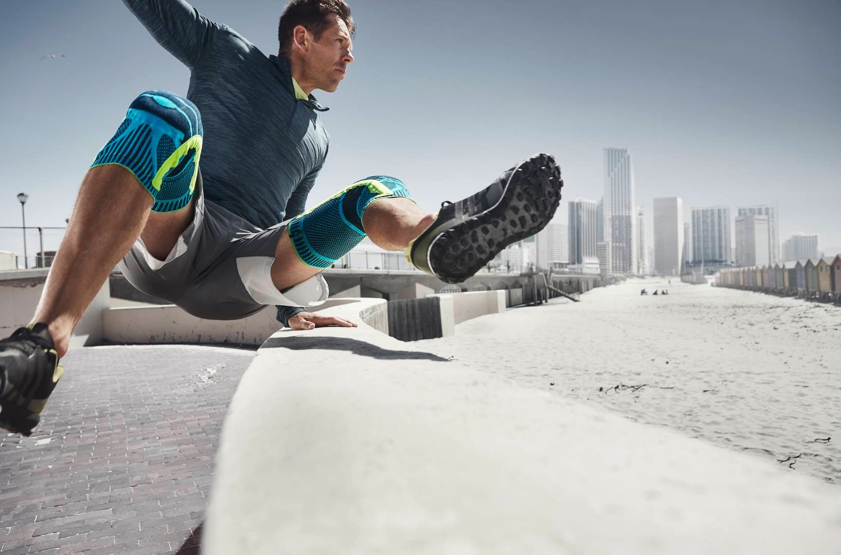 Mann mit zwei Kniebandagen springt beim Urban Running über ein Hindernis	 im Hintergrund Wolkenkratzer