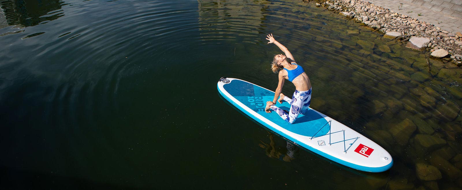 Ein wahrlich schöner Sommertrend: SUP Yoga Frau macht Yoga-Übung auf SUP Board in der Nähe des Ufers	 unter Wasser sind ein paar Steine sichtbar