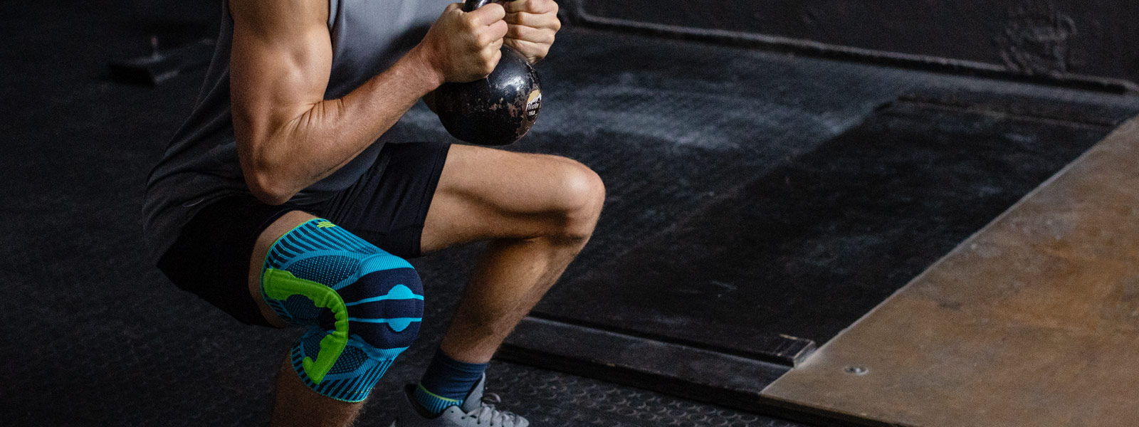 Die Sports Knee Support leistet Dir besonders beim Fitnesstraining großartige Dienste Mann mit Kniebandage geht in die tiefe Hocke mit einer Kettlebell zwischen beiden Händen