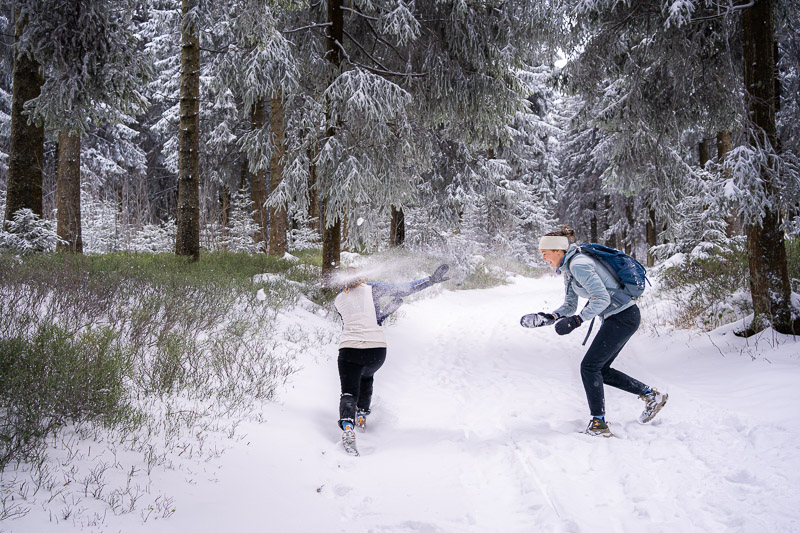 Winterliche Schneeballschlacht – Kindliche Freude im kalten Weiß Zwei Frauen in warmer Winterkleidung haben eine Schneeballschlacht in einem tief verschneiten Wald. Die Frau rechts trägt eine graublaue Jacke und einen Rucksack, während sie gerade einen Schneeball wirft.