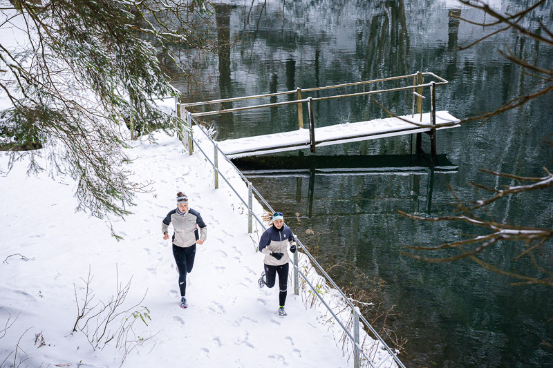 Zwei Läuferinnen joggen entlang eines schneebedeckten Pfades neben einem See mit einem schneebedeckten Steg. Die Wasseroberfläche des Sees spiegelt die umliegenden Bäume wider. Beide Läuferinnen tragen helle Winterlaufjacken und schwarze Hosen.