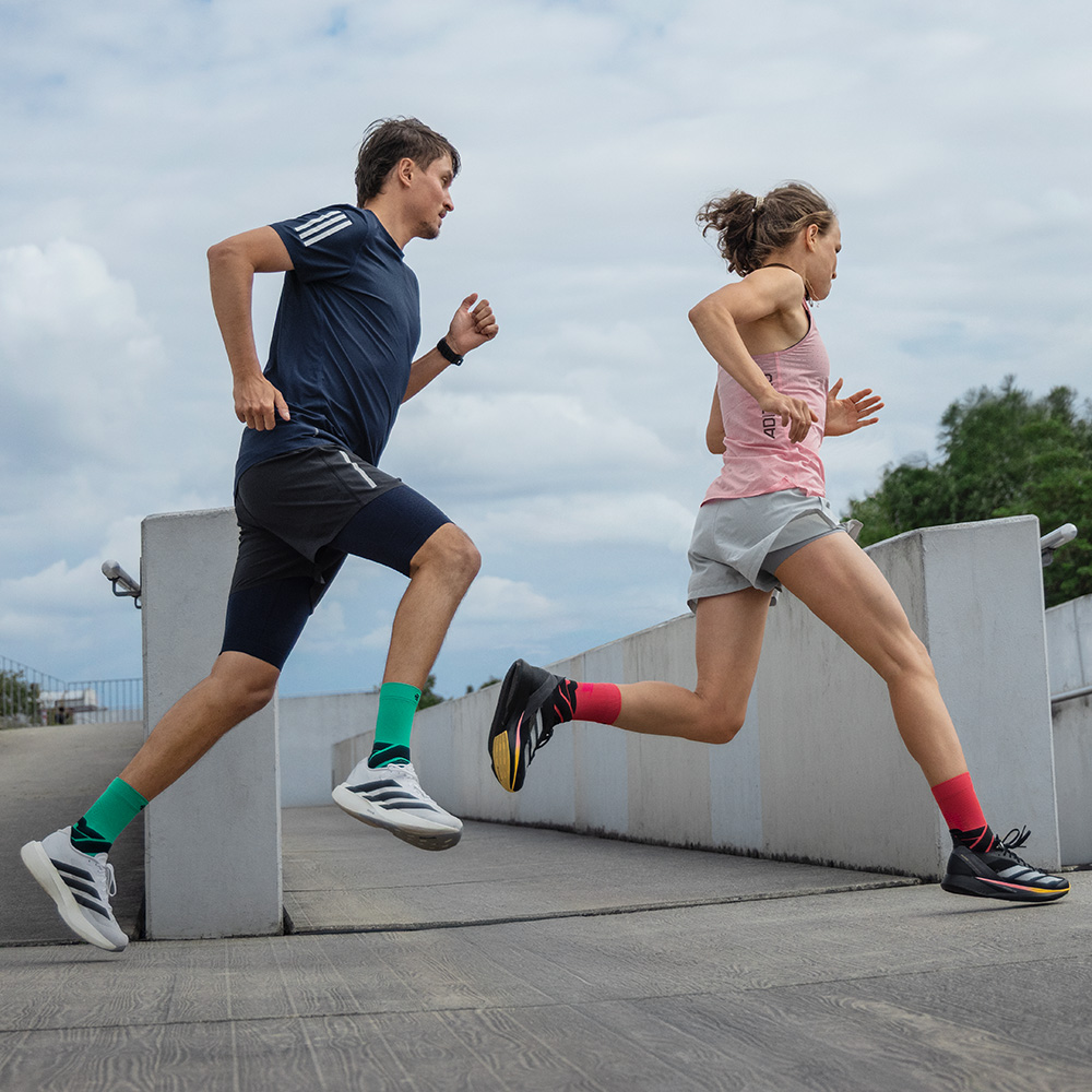 Zwei Läufer in Bewegung auf einer Rampe, ein Mann in blauen Shorts mit grünen Bauerfeind-Laufsocken und eine Frau in grauen Shorts mit roten Bauerfeind-Laufsocken.