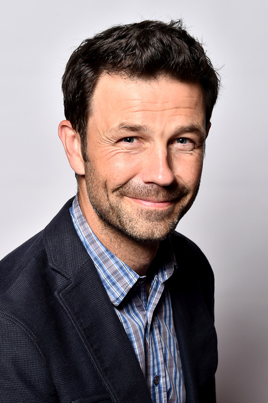 Dr. Casper Grim – Ein freundliches Portrait mit Charisma Ein Mann mit kurzen, dunklen Haaren und einem freundlichen Lächeln trägt ein kariertes Hemd und ein dunkles Sakko. Er blickt direkt in die Kamera vor einem neutralen Hintergrund.
