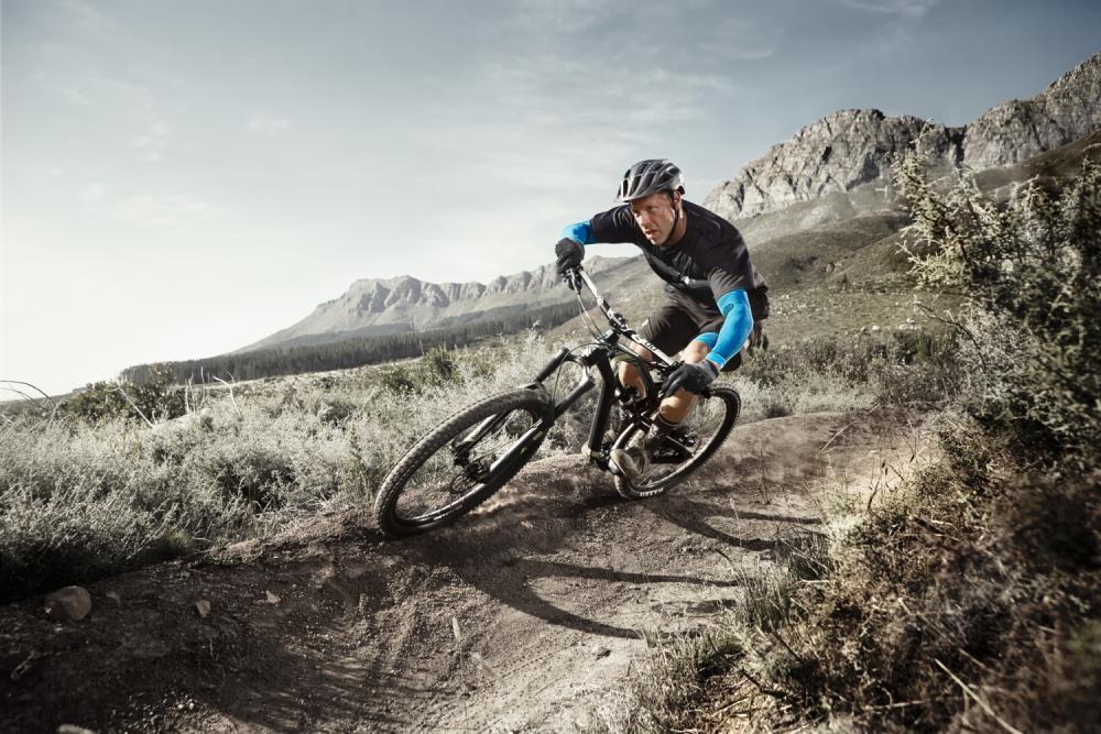 Mann mit Armsleeves fährt auf einem Mountainbike in extremer Kurvenlage auf einem staubigen Weg durch eine karge Landschaft mit Sträuchern und Bergen im Hintergrund