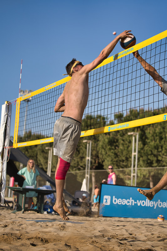Ein Beachvolleyballspieler springt am Netz zum Block, während ein Gegenspieler auf der anderen Seite den Ball schlägt. Der Athlet trägt eine knallrote Kompressionsbandage am rechten Bein.