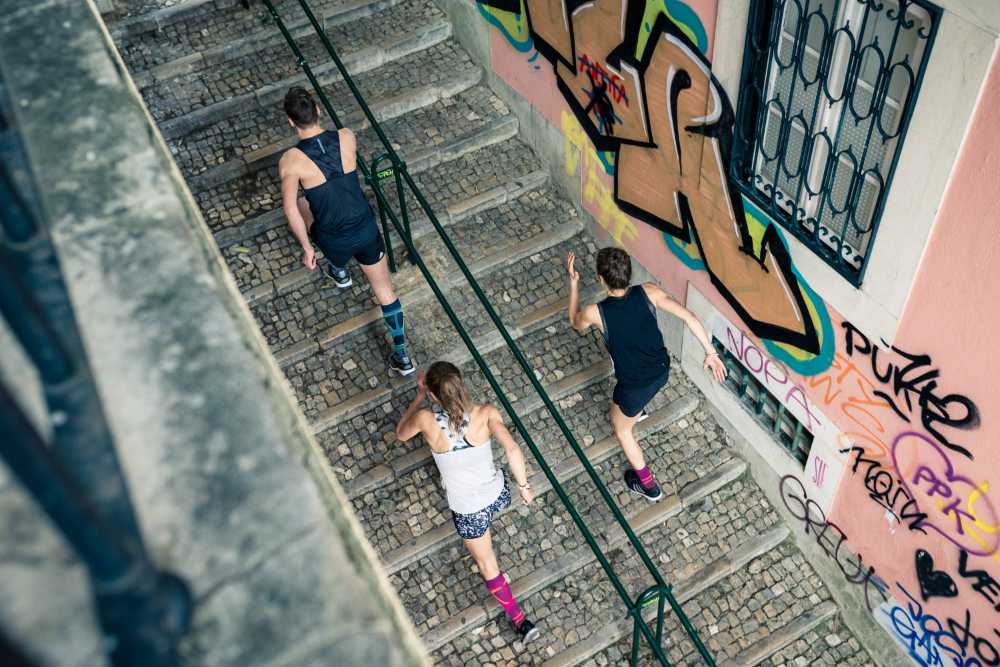 Vogelperspektive: Ein Mann und zwei Frauen in Laufoutfits laufen eine Treppe in einer Altstadt mit Graffitis an den Wänden nach oben
