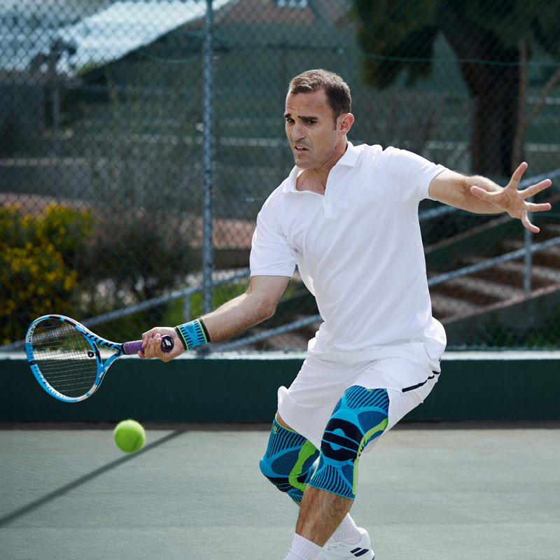 Tennisspieler mit weißer Tenniskleidung und Kniebandagen beim Vorhand Schlag auf einem Hartplatz