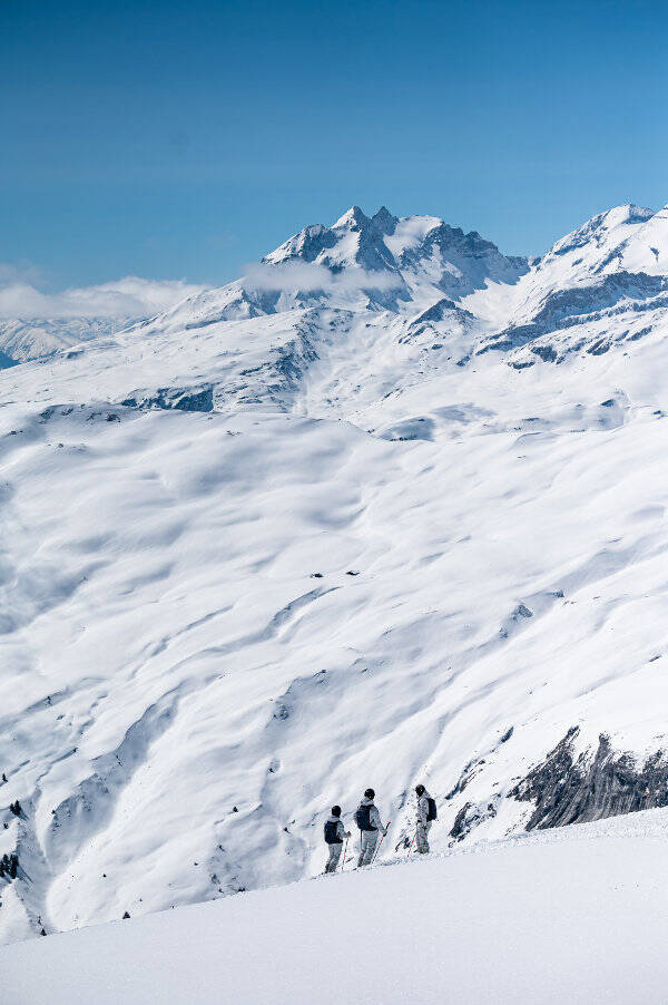 Schneedecktes Alpenpanorama	 davor sind klein drei Skifahrer mit Rucksäcken zu erkennen