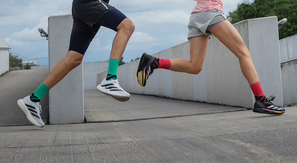 Zwei Läufer in Bewegung, die über eine Rampe sprinten. Der Mann trägt grüne Compression Socks und weiße Laufschuhe, die Frau schwarze Schuhe und rote Compression Socks.