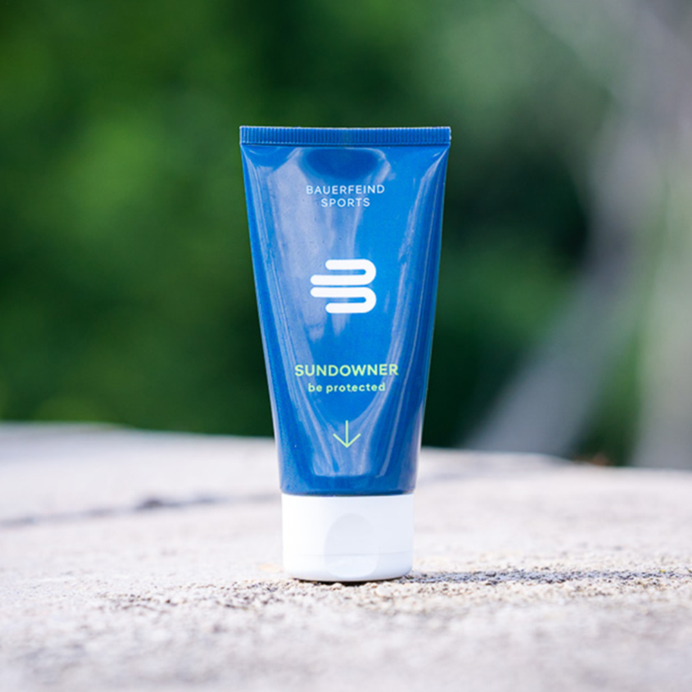 Bauerfeind Sports Sundowner Sonnencreme – UV-Schutz LSF 30 für Outdoor-Aktivitäten Eine blaue Tube der Bauerfeind Sports Sundowner Sonnencreme mit LSF 30 steht auf einem Stein, umgeben von einer grünen Naturkulisse. Die Sonnencreme bietet wasserfesten und veganen Sonnenschutz für Aktivitäten im Freien.