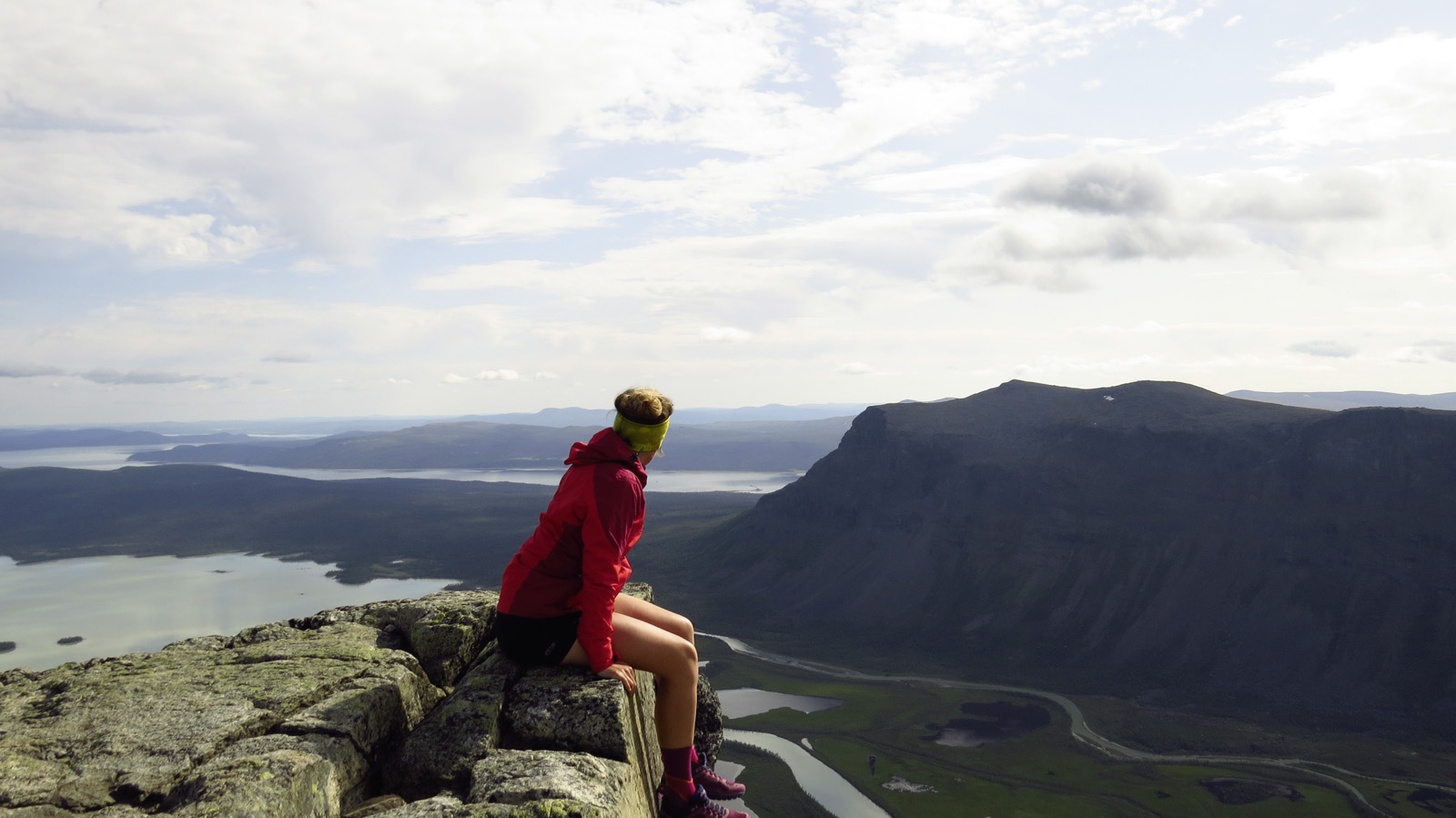 Frau sitzt auf einem Felsvorsprung und schaut in die Ferne in Richtung Berge und Seen