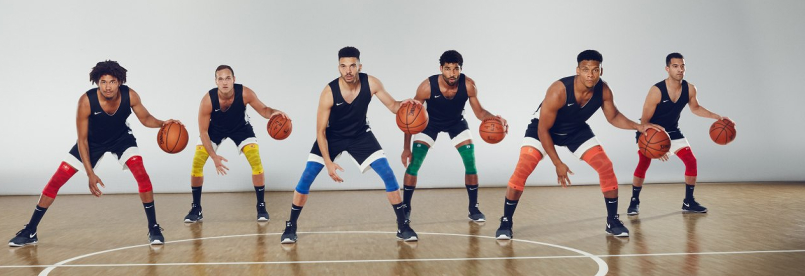 Gemeinsam stark – Fokus und Farbenvielfalt auf dem Court Sechs Basketballspieler stehen in einer Halle auf dem Spielfeld in leicht gebückter Position, jeder mit einem Basketball in der Hand. Sie tragen schwarze Trikots, weiße Shorts und Kompressionsbandagen.