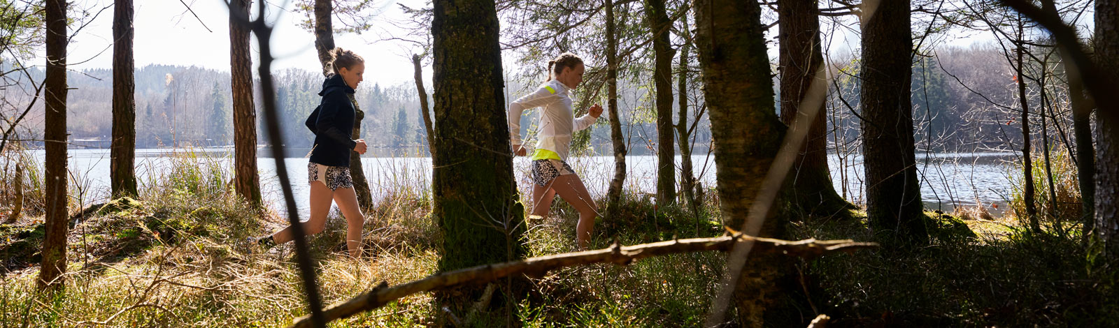 Zwei Läuferinnen laufen hinter Bäumen und vor einem kleinen See hintereinander über einen Waldweg