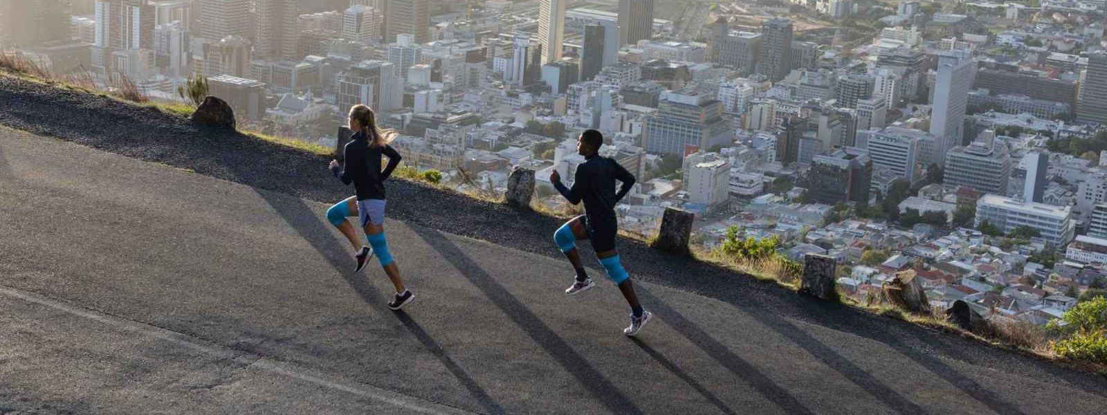 Ganz entspannt oberhalb der Metropole dank der Bauerfeind Sports Compression Knee Support Eine Frau und ein Mann mit blauen Kniebandagen laufen eine Straße oberhalb einer Großstadt hinauf