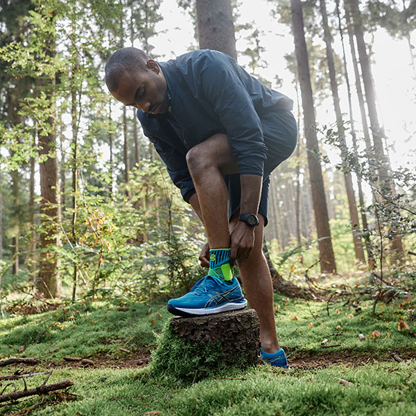 Ein Läufer steht in einem dichten Wald und richtet sich seine Bandage am Knöchel