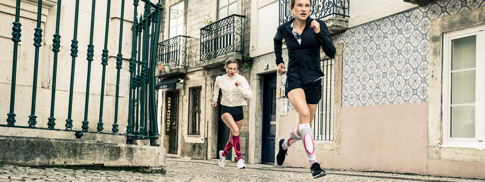 Zwei Frauen rennen energisch durch eine Altstadtstraße mit kleinen Balkonen