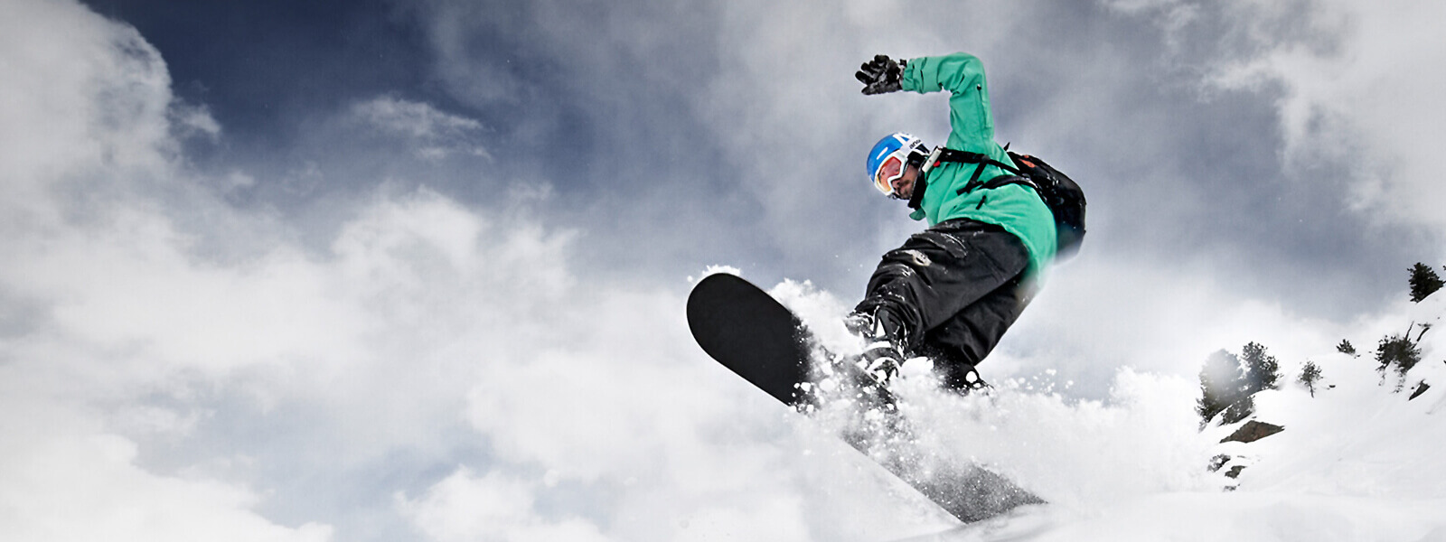Froschperspektive: Snowboarder in grüner Jacke kommt von oben über eine Kuppe gesprungen	 Brettunterseite und viel aufgewirbelter Schnee sind zu sehen