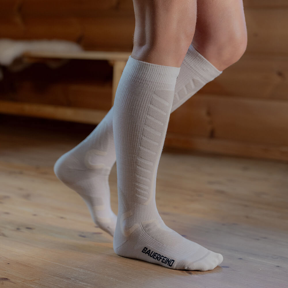 Perfekte Passform: Weiße Kompressions-Skisocken für maximalen Komfort auf der Piste Nahaufnahme von weißen Kompressions-Skisocken, die bis unter die Knie reichen. Die Person steht auf einem Holzboden in einer Hütte. Die Socken bieten durch ihre gepolsterten Zonen zusätzlichen Komfort und Halt beim Skifahren.