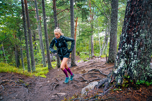 Trailläuferin mit langen Laufsocken und Trainingsjacke läuft einen Waldweg mit vielen Wurzeln hinauf