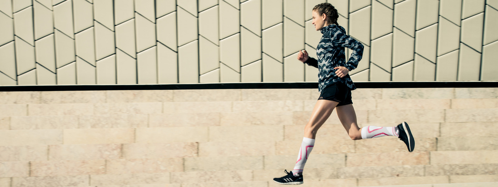 Läuferin mit weiß-rosa Laufsocken läuft neben einer modernen Mauer