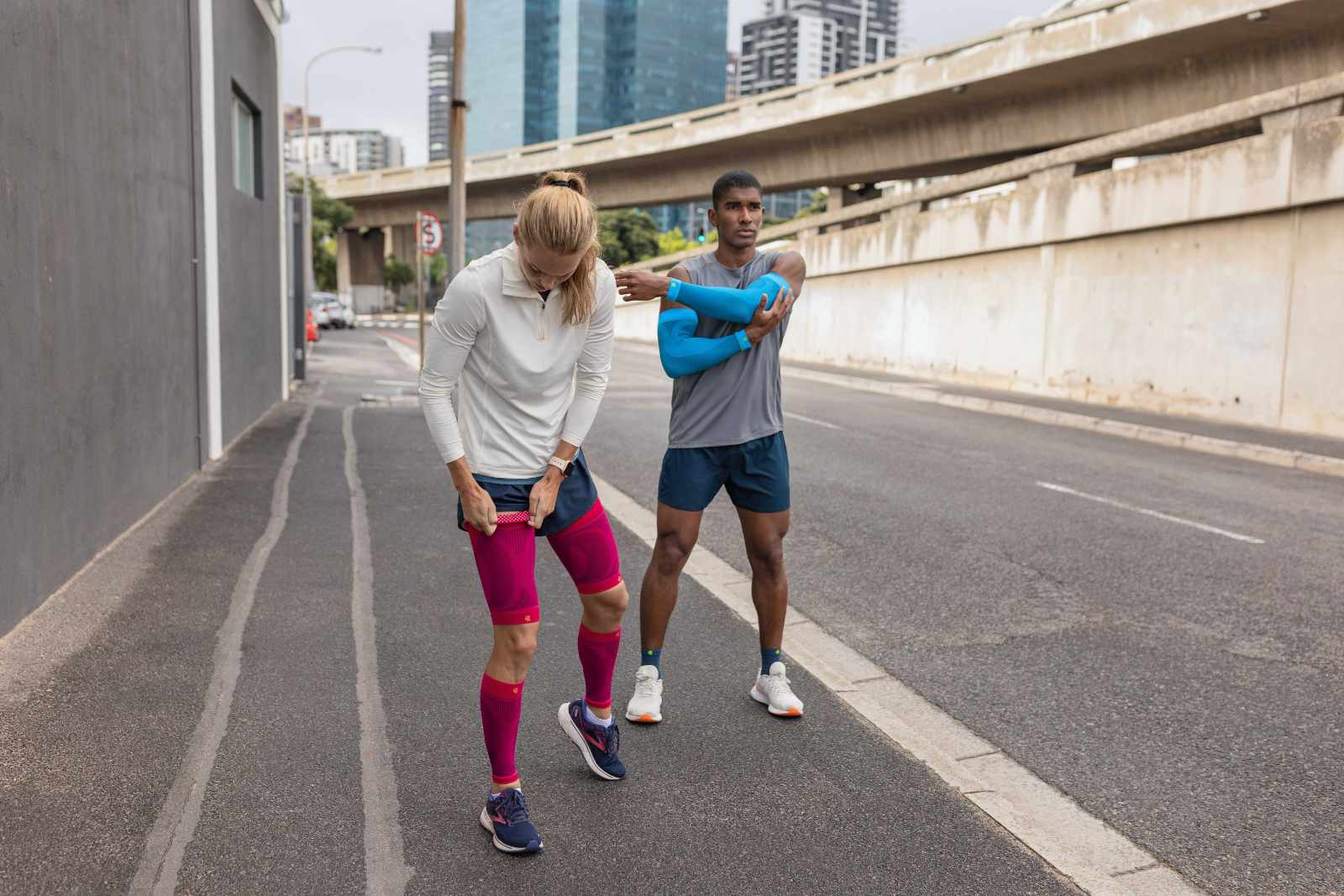 Eine Frau mit pinken Compression Sleeves und ein Mann mit blauen Arm Sleeves und kurzen Laufsocken machen eine Laufpause am Straßenrand einer Großstadt und stretchen sich