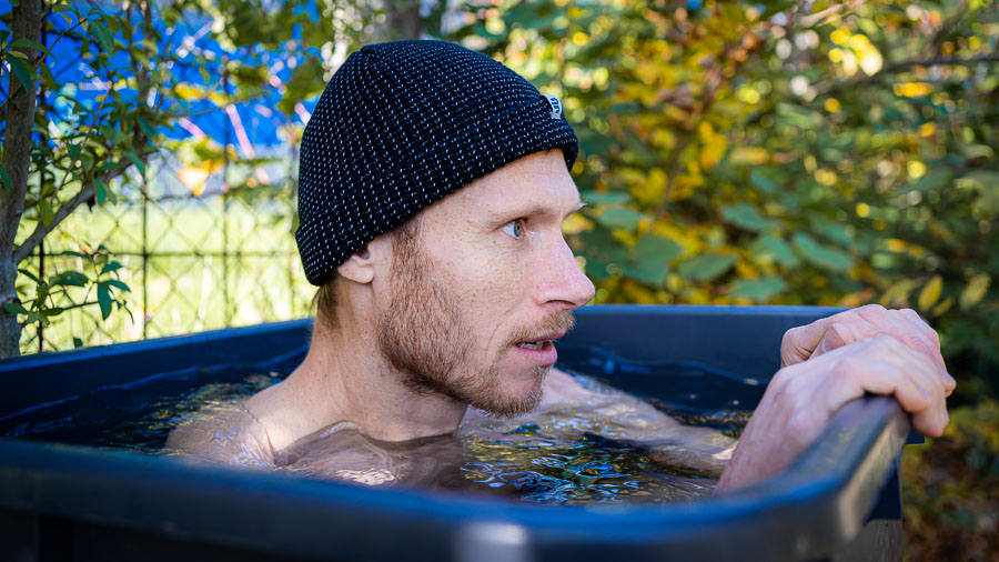 Tief durchatmen: Eisbaden-Challenge in der heimischen Kälte! Ein Mann sitzt in einem schwarzen Wasserbehälter, umgeben von einer herbstlichen Gartenlandschaft. Er trägt eine schwarze Strickmütze und blickt konzentriert zur Seite. Das Wasser reicht ihm bis zur Brust, und der Behälter steht im Freien.