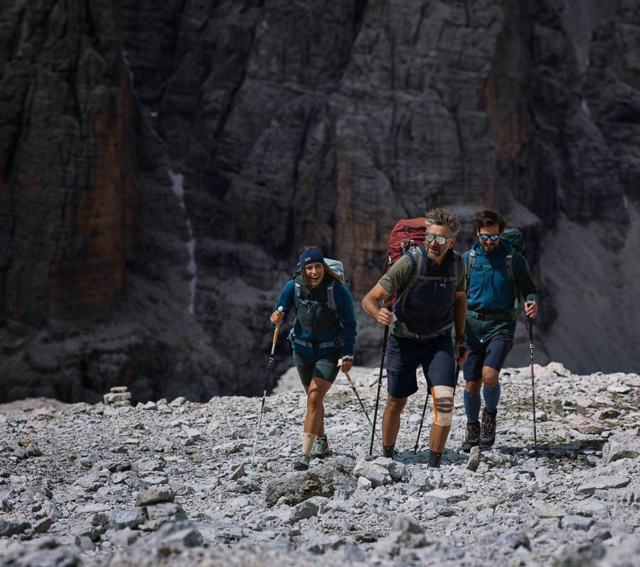 Gemeinsam durch anspruchsvolles Gelände - Wandern mit der richtigen Ausrüstung! Eine Gruppe von drei Wanderern, ausgerüstet mit Rucksäcken und Trekkingstöcken, erklimmt ein felsiges Gelände in den Bergen. Einer der Wanderer trägt eine Kniebandage. Im Hintergrund sind steile Felswände zu sehen.