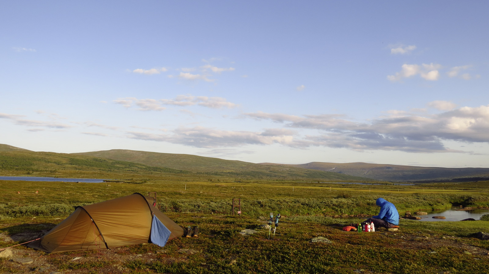 Mann mit Kapuzenjacke sitzt vor einem Zelt in weiter Landschaft vor blauem Himmel