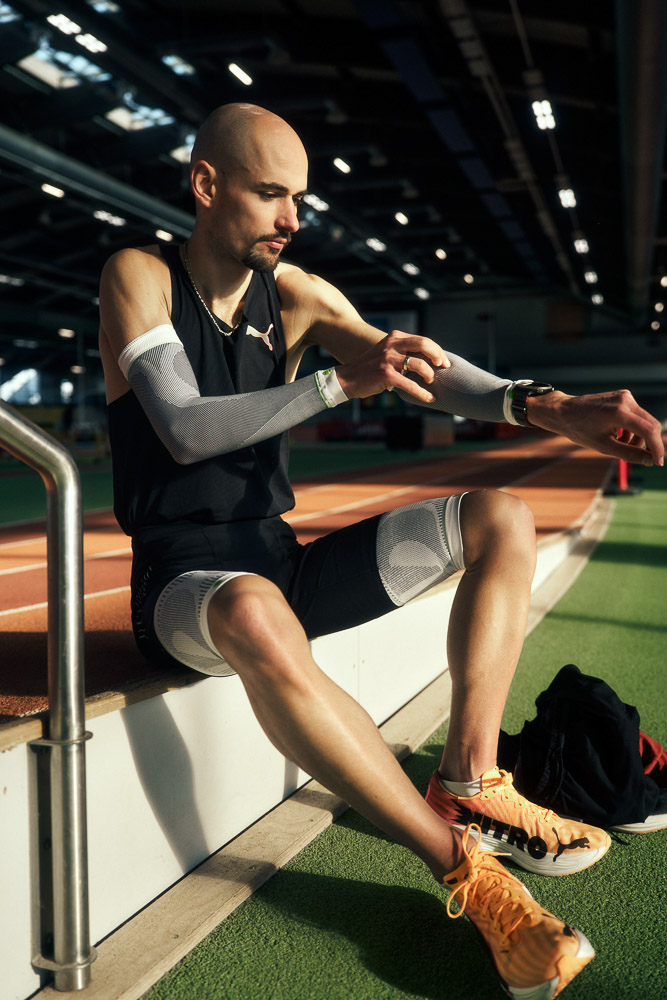 Hendrik Pfeiffer beim Anlegen von Kompressions-Sleeves vor dem Training. Marathonläufer Hendrik Pfeiffer sitzt am Rand einer Indoor-Laufbahn und zieht sich Kompressions-Arm-Sleeves über, als Teil seiner Wettkampfvorbereitung.