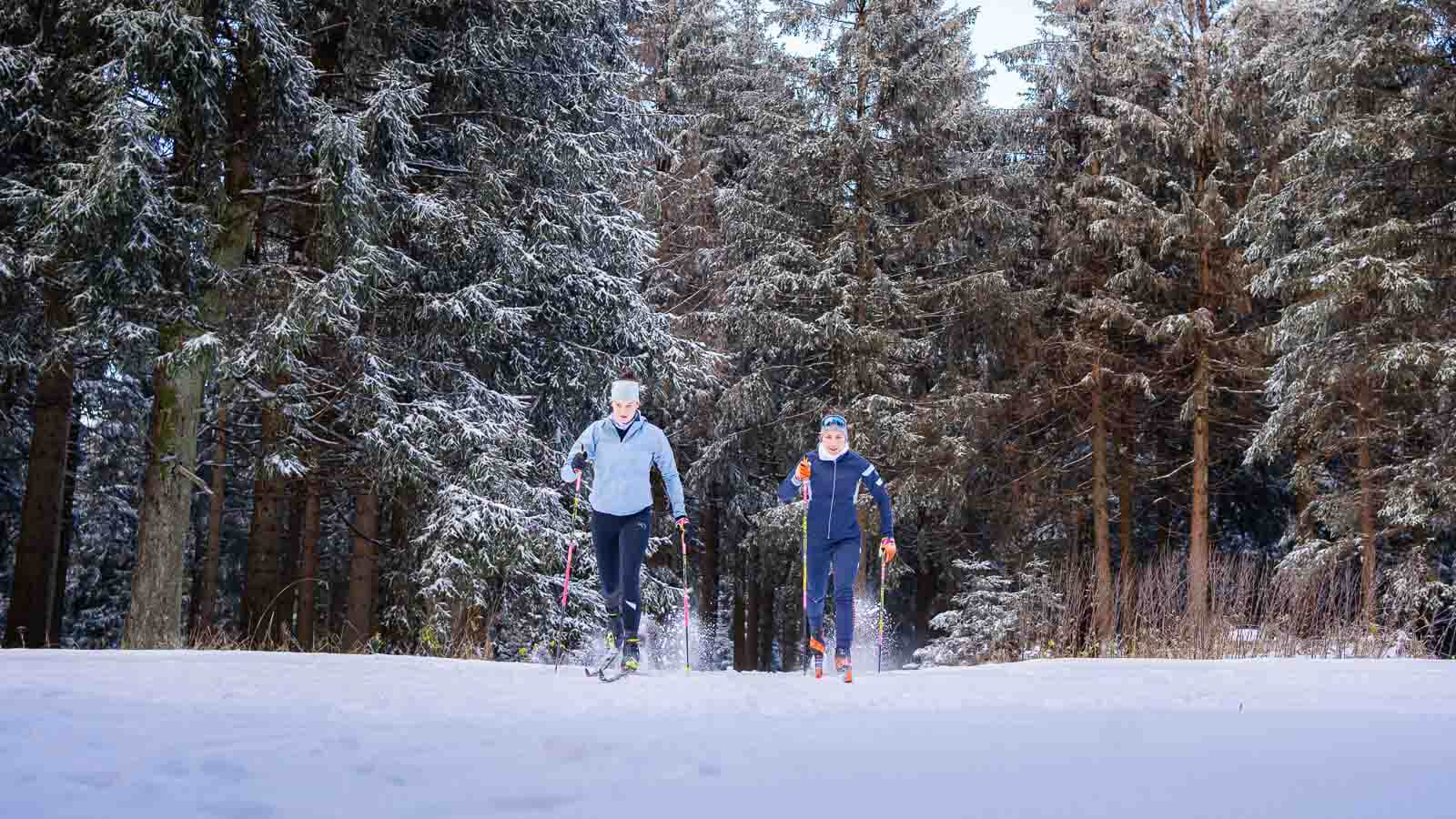 Perfekter Skilanglauf im Winter Zwei Personen laufen beim Skilanglauf durch eine verschneite Landschaft. Beide tragen warme Funktionskleidung, Wintermützen und nutzen Skistöcke. Die Umgebung ist geprägt von verschneiten Bäumen und einer klaren Winteratmosphäre.