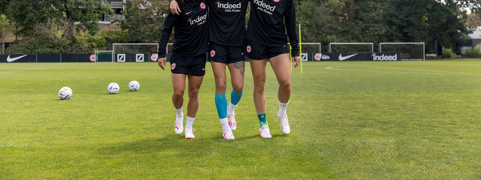 Drei Fußballspielerinnen mit verschiedenen Bandagen umarmen sich auf einen Rasenplatz