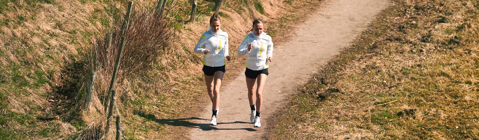 Zwei Läuferinnen in hellen Laufjacken laufen auf einem schmalen Weg zwischen spärlich bewachsenen Wiesen in der Natur