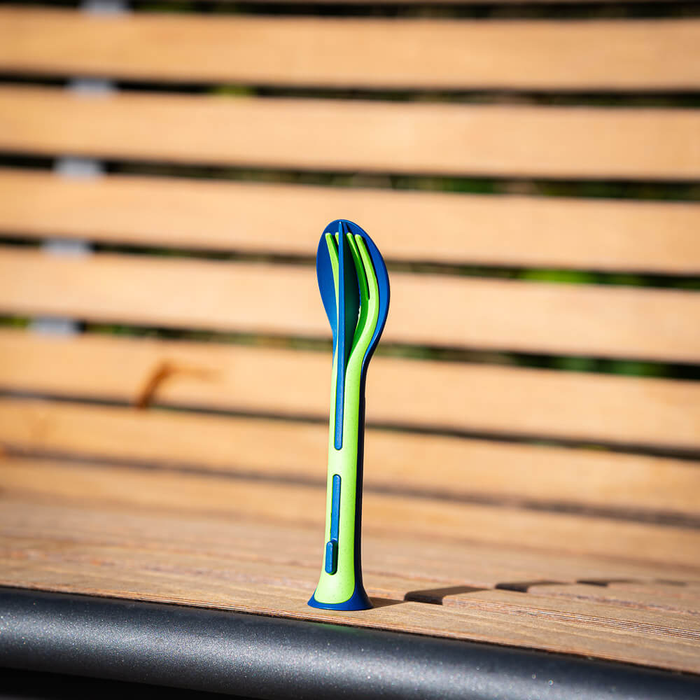 Kompaktes Besteck-Set: Praktisch für Outdoor-Abenteuer Ein grünes und blaues Besteck-Set steht senkrecht auf einer Holzbank. Das Set besteht aus einem Messer, einer Gabel und einem Löffel, die platzsparend ineinander gesteckt sind.