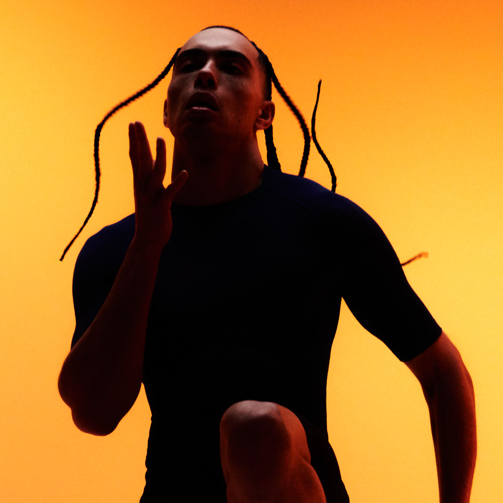 Trainieren mit dem neuen Kompressionskleidung Set für Männer Ein Mann mit Braids macht Kniehebelauf vor orangefarbenen Hintergrund