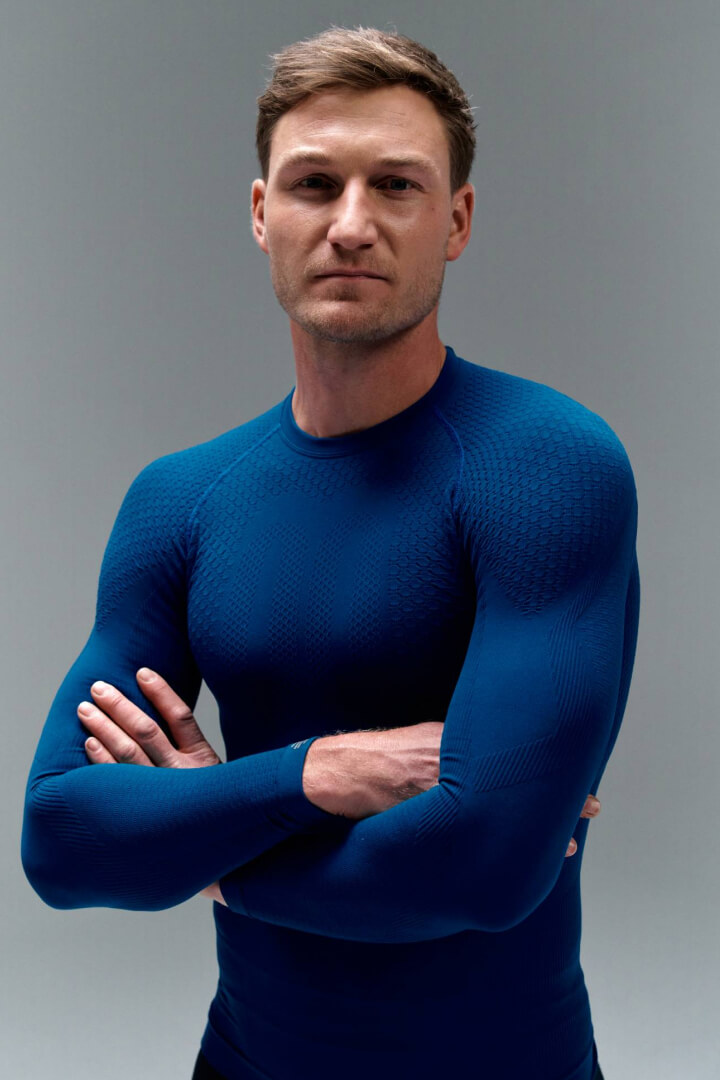 Ein Mann in enganliegendem, funktionalem Sportoberteil in Blau steht mit verschränkten Armen vor grauem Hintergrund. Das Material betont die Muskelstruktur und vermittelt den Eindruck von Kraft, Fokus und Athletik.