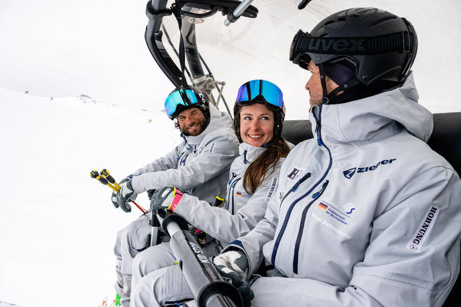Eine Frau und zwei Männer in grauen Skianzügen sitzen gemeinsam in einem Sessellift	 die Frau blickt ihren Kollegen lächelnd an