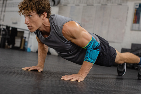 Gerade bei Liegestützen vermeidest Du mit unserer Sports Elbow Support Schmerzen und minderst Belastungsspitzen Junger Mann mit Ellenbogenbandage macht Push-ups auf dem Boden eines Trainingsraums