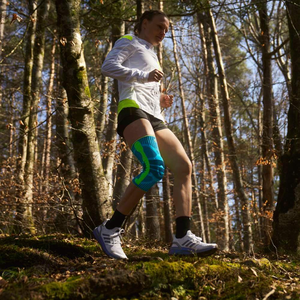 Kniebandage beim Trailrunning im Wald Eine Frau läuft durch einen Waldweg bei Tageslicht, sie trägt eine auffällige türkisfarbene Kniebandage auf dem linken Bein – der Fokus liegt auf Bewegung und Produktnutzung im Outdoor-Kontext.