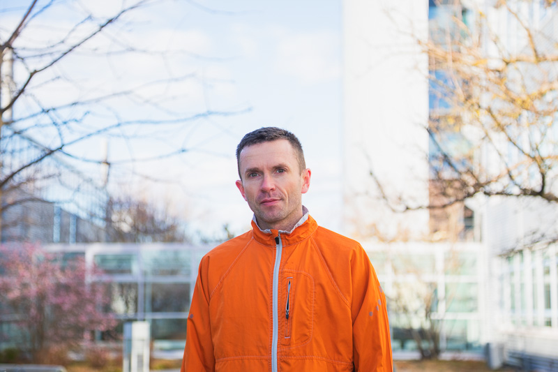 Mit Fokus und Zielstrebigkeit unterwegs Ein Mann in einer leuchtend orangefarbenen Jacke steht vor einem modernen Gebäude und blickt konzentriert in die Kamera. Die kühle Umgebung mit kahlen Bäumen deutet auf die Winterzeit hin.