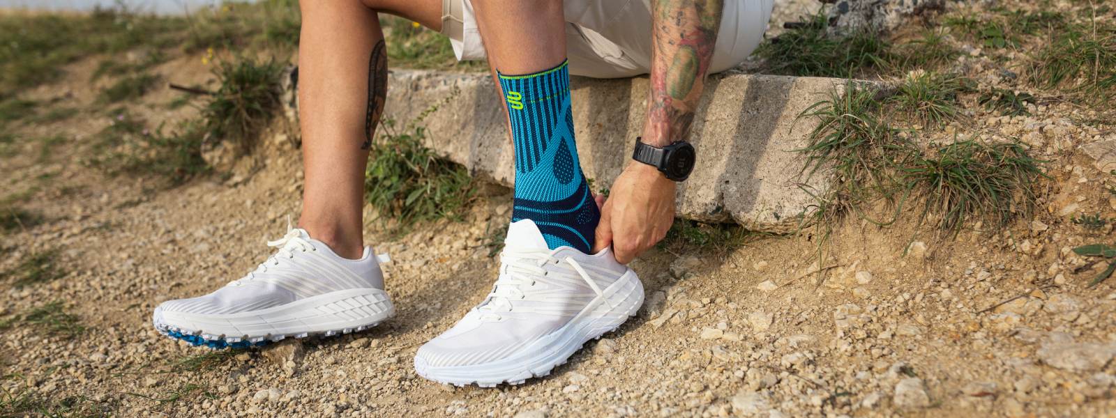 Mit Achillessehnenbandage noch sicherer und schmerzfrei laufen Mann zieht sich auf einem Stein sitzend einen Laufschuh über seine Achillessehnenbandage