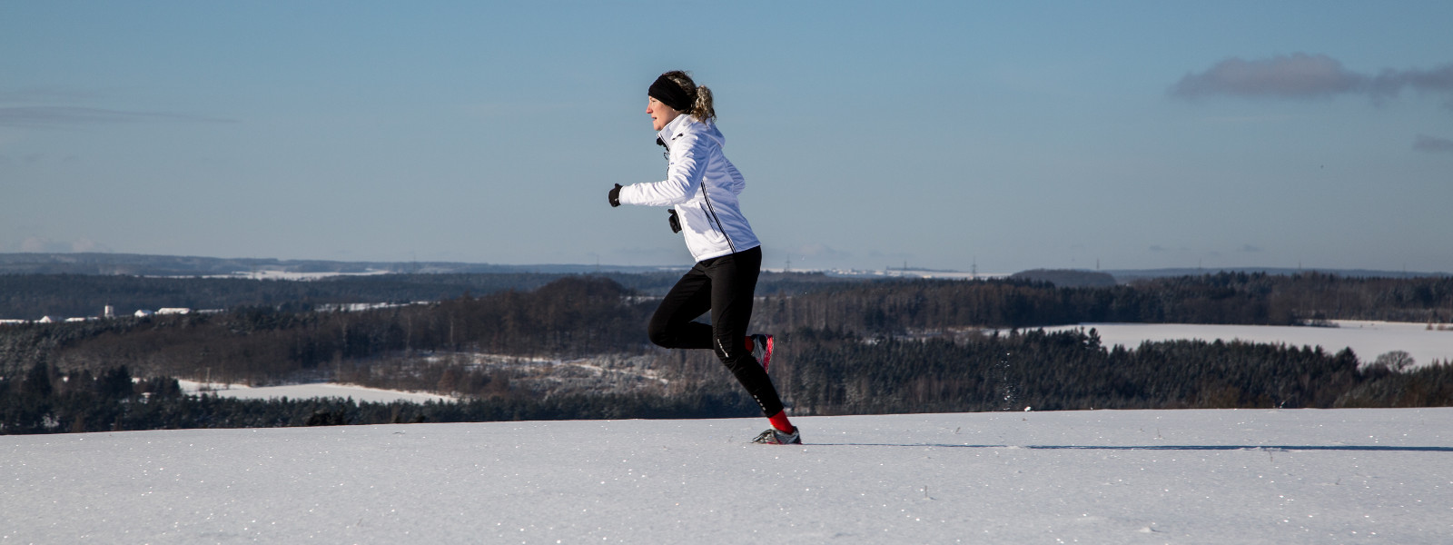 Das Laufen durch Schnee ist nicht nur optisch ein Erlebnis, sondern trainiert noch zusätzlich Deine Muskeln und Gelenke Frau in weißer Laufjacke joggt durch gleichmäßiges Schneefeld im Hintergrund wechseln sich kleine Wälder und Lichtungen ab